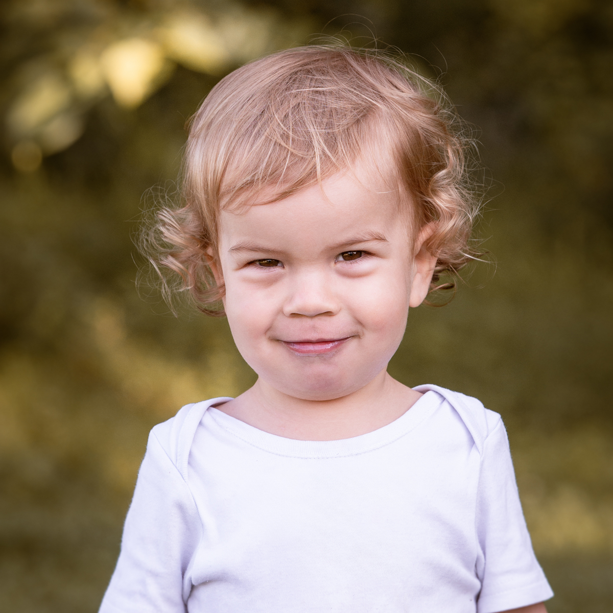 Lächelndes Kleinkind mit lockigem blondem Haar im weißen Shirt vor natürlichem grünem Hintergrund.